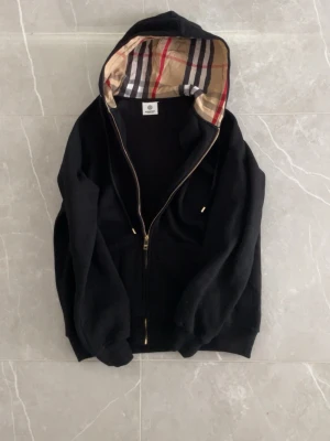 Svart hoodie med Burberry-mönstrad huva - Svart hoodie med dragkedja från Burberry. Huvan är fodrad med det klassiska Burberry-rutiga mönstret i beige, rött och svart. Tröjan har ribbade muddar och fickor framtill. Perfekt för dig som vill ha en stilren och lyxig look. Det är bara att skriva om du har någon fråga 