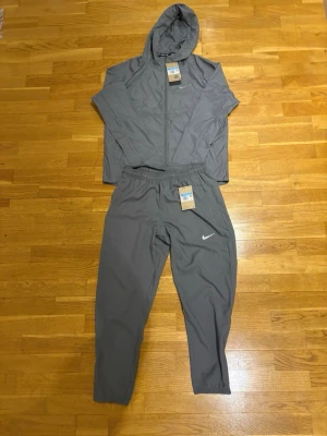 Grått träningsset från Nike - Snyggt grått träningsset från Nike med matchande jacka och byxor. Jackan har huva, dragkedja och diskret Nike-logga på bröstet. Byxorna har resår i midjan och Nike-logga på benet. Perfekt för träning eller chill. Tillverkat i lätt och funktionellt material.