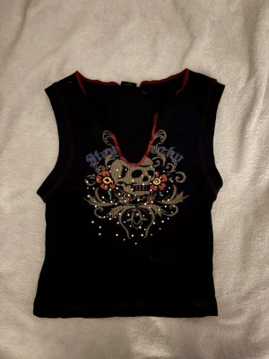 Svart ärmlös topp med döskalletryck - Säljer en svart ärmlös topp med coolt dödskalle tryck med blommor och stenar. Toppen har röda kontrastsömmar och en croppad passform💗
