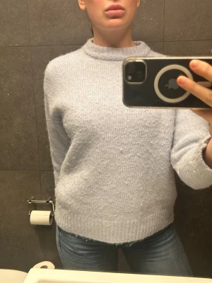 Ljusblå stickad tröja med rund hals - Mysig ljusblå stickad tröja med rund hals och långa ärmar. Tröjan har en seating sopass loose fit och ribbstickade mud gör den apart. Perfekt för lager på lager och chill taller. Materialet ser ut att vara mjukt och fluffigt, troligen ullblandning.