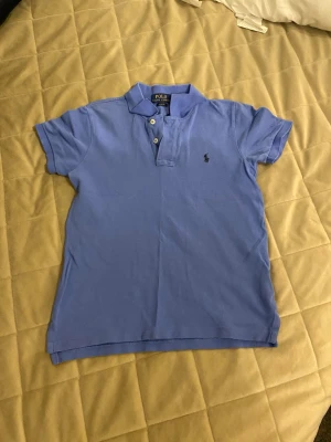 Blå piké från Polo Ralph Lauren - Klassisk blå pikétröja från Polo Ralph Lauren med korta ärmar och krage. Tröjan har två knappar vid halsen och den ikoniska lilla logotypen broderad på bröstet. Perfekt till sommaren. OBS! Tröjan är solblekt och har en liten defekt av lappen som sitter i tröjan som syns på baksidan. (Bild 4) Inget som syns på håll.