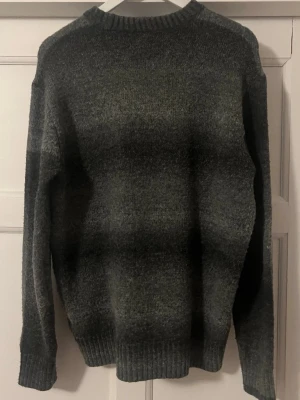 Mörkgrå stickad tröja med tonade ränder - Mysig stickad tröja i mörkgrått med subtila tonade ränder. Tröjan har rund halsringning, långa ärmar och ribbstickade muddar vid ärmslut och nederkant. Perfekt för kyliga dagar och enkel att matcha med jeans eller kjol.