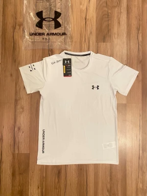 Vit Under Armour t-shirt - Snygg vit t-shirt från Under Armour med svart logga på bröstet och tryck på ärmen. T-shirten är kortärmad och har en sportig passform, perfekt för träning. Materialet är lätt och andas, vilket gör den bekväm att ha på sig under aktiva dagar.