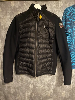 Svart pufferjacka från Parajumpers - Svart pufferjacka från Parajumpers med quiltad front och rygg, stickade ärmar och axlar. Jackan har hög krage, gul detalj vid dragkedjan och en bröstficka med dragkedja. Märkespatch på vänster ärm. Perfekt för kalla dagar. Funkar även på våren. Jätteskönt jacka för dig som vill ha lite skön stil 