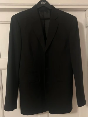 Svart kavaj från Giorgio Armani - Stilren svart kavaj från Giorgio Armani med klassisk passform och snygga slag. Kavajen har två fickor framtill och är helfodrad med elegant svart innerfoder. Perfekt för en dressad look och passar till många olika tillfällen.