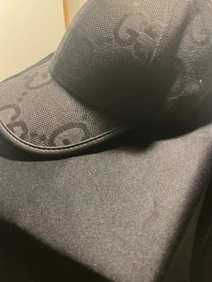 Svart keps från Gucci med GG-mönster - Snygg svart keps från Gucci med det klassiska GG-mönstret i ton-i-ton. Kepsen har justerbart spänne bak i läder och detaljer i guld. Tillverkad i canvasmaterial med läderdetaljer för en exklusiv känsla.