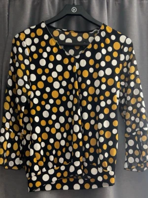 Prickig topp med trekvartsärm - Snygg topp med svart bas och stora vita och gula prickar. Toppen har trekvartsärm och v-ringad hals. Materialet känns mjukt och stretchigt, troligen polyester. Perfekt för dig som gillar färg och mönster!