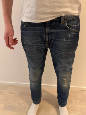 Grim Tim jeans från Nudie Jeans - Säljer ett par blå slim fit jeans från Nudie Jeans med slitningar och tvättade detaljer. Klassisk femficksmodell med orange sömmar och broderade bakfickor. Jeansen har normal midja och är tillverkade i denim av bomull. Modell Grim Tim Yesterdays News. Inte längre till salu. Nypris 2000kr storlek W30 L30