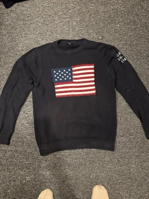 Mörkblå stickad tröja Gant USA-flagga - Snygg mörkblå stickad tröja från Gant med stor amerikansk flagga på bröstet. Rund halsringning och ribbade muddar. Diskret Gant-brodyr på ärmen. Perfekt för dig som gillar klassisk och sportig stil.