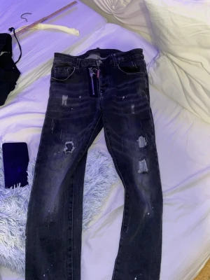 Dsquared2 jeans  - Säljer ett par svarta jeans med slitningar och distressed detaljer. Jeansen har en unik dragkedja med ring framtill och klassisk femficksdesign. Perfekta för dig som gillar en edgy och avslappnad stil.