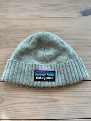 Grå mössa från Patagonia - Snygg grå mössa från Patagonia med ribbstickad kant och klassisk logotyp framtill. Tillverkad i mjukt stickat material som håller dig varm. Enkel och stilren design som passar till det mesta.