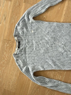 Ljusblå stickad tröja från Gina Tricot - Ljusblå stickad tröja från Gina Tricot med kabelstickat mönster och diskret logga broderad på bröstet. Tröjan har rund halsringning, långa ärmar och ribbade muddar vid ärmslut och nederkant. Perfekt för en chill och stilren look.