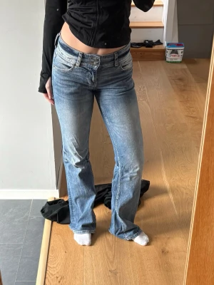 Blå bootcut jeans med låg midja - Säljer ett par blå bootcut jeans med låg midja och klassisk femficksmodell. Jeansen har en snygg tvätt med slitningar framtill och lätt utsvängda ben. Perfekta till sneakers eller boots för en avslappnad look.