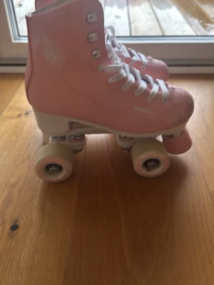 Rosa rullskridskor från Oxelo - Säljer ett par klassiska rosa rullskridskor från Oxelo med vita snören och vita hjul. Skorna har hög modell och tåskydd framtill. Perfekta för dig som vill cruisa runt med stil på gatorna eller i parken.