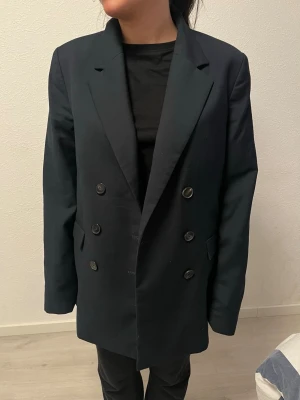 Svart dubbelknäppt oversized kavaj - Stilren svart kavaj med dubbelknäppning och breda slag. Oversized passform som ger en trendig look. Fyra knappar på varje sida framtill och två fickor. Perfekt att styla med både jeans och kostymbyxor för en cool vibe.