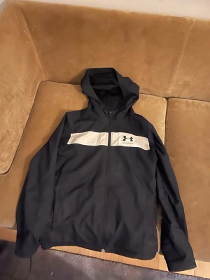  Under Armour vindjacka  - Vindjacka från Under Armour med dragkedja framtill och en vit panel över bröstet där loggan syns. Tillverkad i ett mjukt och sportigt material, perfekt för chill eller träning. Klassisk huva och långa ärmar ger en clean och sportig look.