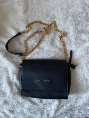 Svart axelväska från Valentino - Snygg svart axelväska från Valentino med guldfärgad kedjerem och stilren design. Väskan har ett lock med diskret logga framtill och detaljer i guld. Tillverkad i slätt konstläder och har en justerbar axelrem för extra komfort.