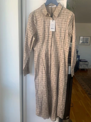 Beige rutig långklänning från NA-KD - Stilren beige långklänning från NA-KD med svart rutigt mönster. Klänningen har krage, knäppning framtill och långa ärmar. Perfekt för dig som gillar minimalistisk och modern stil.