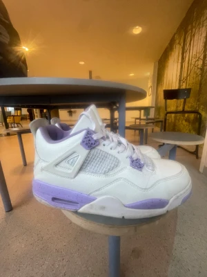 Vita och lila Air Jordan 4 sneakers - Snygga Air Jordan 4 sneakers i vitt läder med lila detaljer på snörning, sula och innersida. Klassisk meshpanel på sidorna och ikoniska plastdetaljer. Perfekta för dig som vill sticka ut med stilrena och trendiga sneakers.