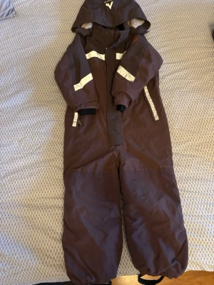 Bruna täckbyxor med huva McKinley - Bruna täckbyxor från McKinley med huva och reflexdetaljer på armar och bröst. Dragkedja framtill och resår vid ärmslut och benslut. Perfekt för kalla dagar och utomhuslek. Slitstarka och vattenavvisande material.