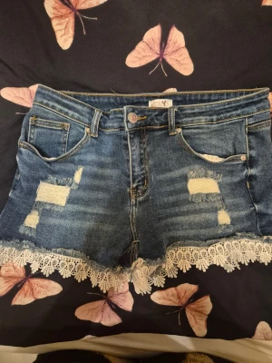 Blå jeansshorts med spets från Hailys - Säljer ett par blå jeansshorts från Hailys med slitna detaljer och vit spetskant längst ner. Shortsen har klassisk femficksdesign, knappgylf och råa kanter för en trendig look. Perfekta för varma dagar och ger en cool vibe.