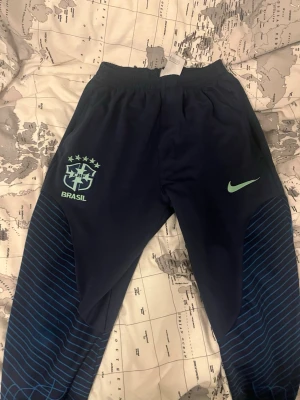 Nike Brasil Dri-FIT träningsbyxor - Mörkblå träningsbyxor från Nike med Brasil-logga och Dri-FIT-material. Byxorna har elastisk midja, ljusgrönt Nike-märke och Brasil-tryck på ena benet samt blåa linjemönster på benen. Perfekta för fotbollsträning eller gymmet.