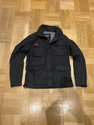 Superdry vårjacka  - Säljer en snygg och praktisk Superdry field jacka i mörkgrön färg. Jackan är i storlek L men passar bättre till dig som vanligtvis har M.  ✔ Mycket bra skick ✔ Klassisk field jacket-modell ✔ Flera fickor och robust material ✔ Perfekt för höst, vår och mild vinter