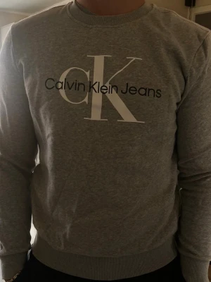 Grå sweatshirt från Calvin Klein - Snygg grå sweatshirt från Calvin Klein Jeans med stort CK-tryck och logga på bröstet. Tröjan har rund halsringning, ribbade muddar vid ärmslut och nederkant. Perfekt för en avslappnad och trendig look.