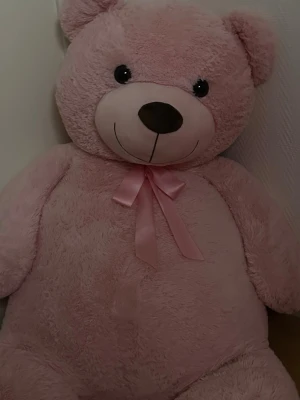 Stor rosa nallebjörn med rosett från Ullared  - Mysig och stor rosa nallebjörn med mjuk plysch och en söt rosa rosett runt halsen. Nallen har stora svarta ögon, rund nos och ett gulligt leende. Perfekt som present eller dekoration i rummet.