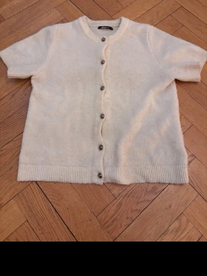 Krämvit kortärmad kofta - Kortärmad kofta i mjukt, krämvitt material med rund hals och ribbade kanter. Knäpps med mörka knappar framtill. Perfekt att slänga över en topp för en chill look. Frågor?? Kontakta mej