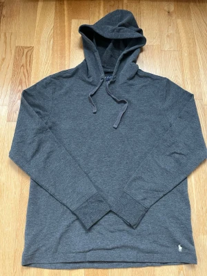 Grå hoodie från Polo Ralph Lauren - Mysig grå hoodie från Polo Ralph Lauren i mjukt material, perfekt för chill dagar hemma. Tröjan har huva med dragsko, långa ärmar och en liten broderad logga vid nederkanten. Enkel och stilren design som passar perfekt till loungewear.
