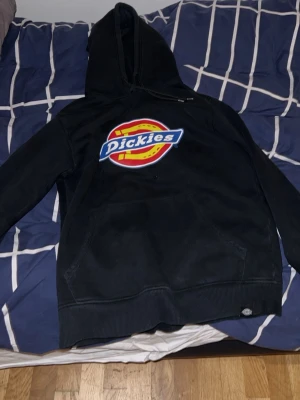 Svart Dickies hoodie med stor logga - Säljer en svart hoodie från Dickies med stor färgglad logga på bröstet. Klassisk känguruficka framtill och huva med dragsko. Perfekt för dig som gillar streetwear och vill ha en bekväm och stilren hoodie.