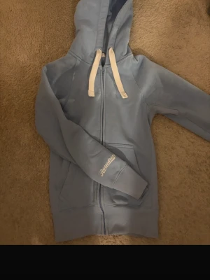 Ljusblå hoodie från Russemerch  - Ljusblå hoodie med dragkedja från Russemerch Tröjan har huva med vita snören, två fickor framtill och broderad logga på ärmen. Tillverkad i mjukt material som känns skönt mot huden. Perfekt för en avslappnad och stilren look.