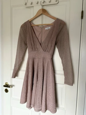 Glittrig champagnefärgad klänning Bubbleroom - Supersnygg glittrig kortklänning från Bubbleroom i rosa champagnefärg. Klänningen har v-ringning, långa ärmar och ett medföljande bälte som knyts i midjan. Perfekt för dig som vill sticka ut med lite extra glitter.