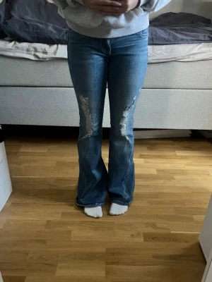 Blå Gina tricot bootcut jeans med slitningar - Använda två gånger, Säljer ett par blå Gina  bootcut jeans med tydliga slitningar på låren och knäna. Jeansen har en klassisk femficksmodell och är i ett stretchigt denimtyg. Perfekta för dig som gillar en avslappnad och trendig look.