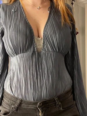 Grå plisserad body med v-ringning - Snygg grå body med plisserat tyg och djup v-ringning. Långa ärmar och figurnära passform ger en trendig look. Materialet är glansigt och stretchigt, vilket gör den bekväm att bära. Perfekt att styla med jeans eller kjol för en cool outfit. Färgen är en blandning av blå,grön 