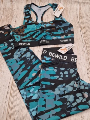 Träningsset från Bewild i turkos camo - Snyggt träningsset från Bewild med sport-bh och tights i blått, svart och turkost camomönster. Setet har breda resårband med logga och är gjort i stretchigt funktionsmaterial som passar perfekt till gymmet eller löpning.