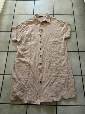 Beige/apricos skjortklänning med knappar, bomull  - Luftig skjortklänning i bomull med korta ärmar och knappar hela vägen framtill. Klänningen har två stora fickor fram och en klassisk krage. Perfekt för varma dagar och enkel att styla.