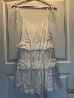 Vit volangklänning med knytband  - Söt vit klänning med volanger och tunna axelband som knyts upptill. Klänningen har flera lager volanger och är tillverkad i ett lätt, luftigt material som passar perfekt way varma dagar. Den har en avslappnad passform och är ärmlös. (Den behövs strykas) 