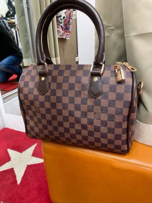 Marcus handväska brun - Säljer en klassisk väska i brun  Väskan har två robusta handtag i mörkbrunt läder, guldfärgade metalldetaljer och dragkedja. Tillverkad i canvas med läderdetaljer och ikoniskt rutmönster.