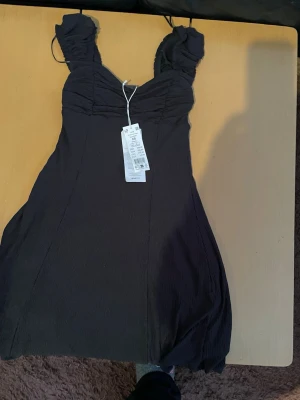 Svart kort klänning med volang - Snygg svart kort klänning med volangdetaljer vid axelbanden och rynkad byst. Klänningen har en feminin siluett och tunna axelband. Perfekt för dig som vill ha en enkel men elegant look.