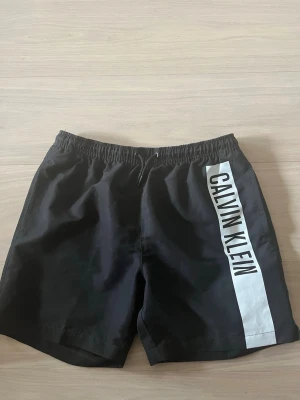 Svarta badbyxor Calvin Klein - Snygga svarta badbyxor från Calvin Klein med vit sidopanel och stor logga längs benet. Klassisk modell med resår i midjan och vit mesh på insidan för extra komfort. Perfekta för stranden eller poolen.