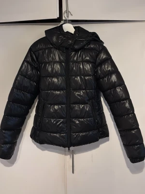 Svart pufferjacka med huva - Säljer en svart pufferjacka med huva och dragkedja framtill. Jackan har en glansig finish, quiltad design och två sidofickor. Perfekt för kalla vinterdagar och riktigt skön att bära. Materialet är troligtvis syntetiskt och jackan har en modern, avslappnad passform.