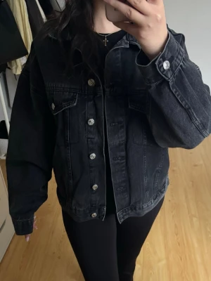 Svart oversized jeansjacka - Snygg svart jeansjacka i oversized modell med klassiska silverfärgade knappar och två bröstfickor med lock. Endast använt vid ett tillfälle.