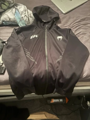 Svart UFC hoodie från Venum - Svart hoodie från Venum med UFC-logga på bröstet och dragkedja framtill. Tillverkad i 94% polyester och 6% elastan för en skön och stretchig känsla. Snygga detaljer på ärmarna och huva för extra stilpoäng.