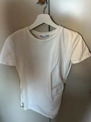 Vit t-shirt med knyt från Zara - Säljer en vit t-shirt från Zara. T-shirten har korta ärmar, rund halsringning och en snygg knyt-detalj i sidan 