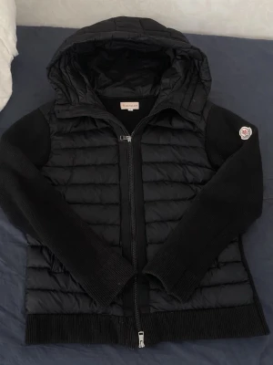 Moncler cardigan - Snygg svart pufferjacka från Moncler med stickade ärmar och huva. Jackan har quiltad front och rygg i blankt material, dragkedja framtill och Moncler-logga på ärmen. Perfekt för kalla dagar och riktigt trendig look.