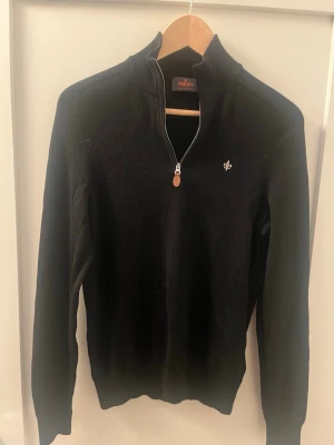 Svart halvzip-tröja från Morris - Stilren svart tröja från Morris med halv dragkedja och liten broderad logga på bröstet. Tröjan har hög krage och ribbade muddar vid ärmslut och nederkant. Perfekt för en clean och klassisk look.