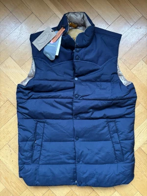 Marinblå Primaloft väst från Rocky Mountain Featherbed - helt ny - L - Snygg marinblå dunväst från Rocky Mountain Featherbed med hög krage och tryckknappar framtill. Västen har två stora fickor och beige insida. Tillverkad i återvunnen nylon och dunfyllning för extra värme. Perfekt för lager-på-lager och streetwear.