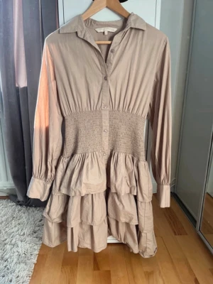 Beige volangklänning med knappar - Beige kortklänning med volanger och knappar framtill. Klänningen har lång ärm, markerad midja med smock och klassisk krage. Tillverkad i mjukt bomullsmaterial och har en avslappnad, men feminin siluett.